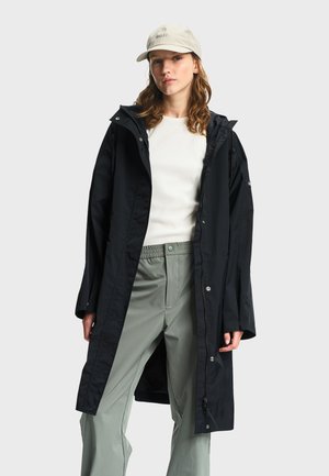 Femme portant une casquette beige, une chemise blanche, un long manteau noir et un pantalon gris clair, debout devant un fond clair uni.