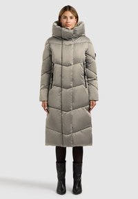 Gepolsterter langer Mantel in Hellgrau mit hohem Kragen, gestepptem Design, seitlichen Reißverschlusstaschen und geradem Schnitt. Kombiniert mit schwarzen Stiefeln.