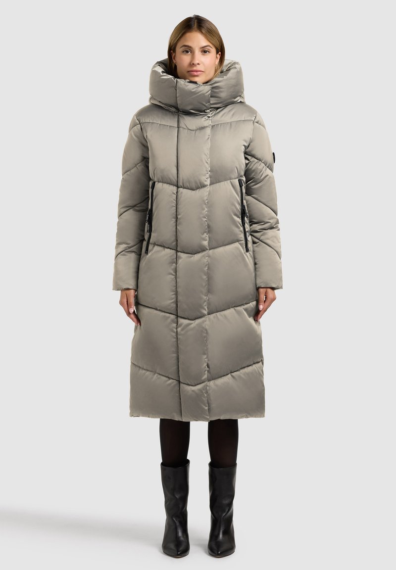 Gepolsterter langer Mantel in Hellgrau mit hohem Kragen, gestepptem Design, seitlichen Reißverschlusstaschen und geradem Schnitt. Kombiniert mit schwarzen Stiefeln.