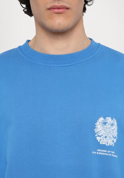 Blaues Baumwoll-Sweatshirt mit geripptem Ausschnitt, featuring einem weißen Adler-Logo und Text, glatte Textur und lockere Passform.