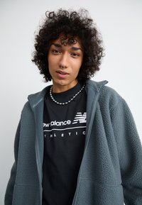 Šedá fleecová bunda na zip přes černé tričko se bílým nápisem "New Balance Athletic". Hladká tkanina s volným střihem, detailní pohled.