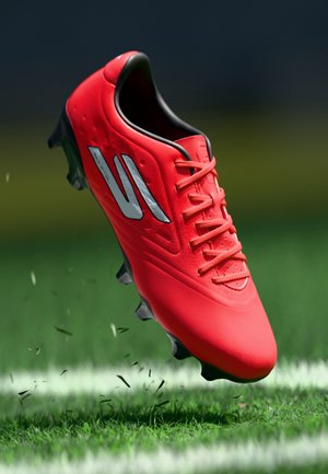 Leuchtend roter Fußballschuh mit schwarzen Stollen, der Gras auf einem markierten grünen Feld hochwirbelt, in der Luft aus einem Winkel gezeigt.