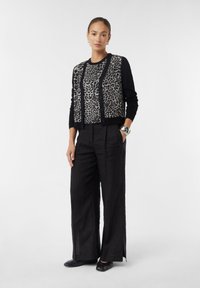 Schwarze Strickjacke mit Leopardenmuster auf der Vorderseite, Knopfverschluss und langen Ärmeln. Kombiniert mit lockeren schwarzen Hosen und schwarzen Schuhen. Silbere Uhr am Handgelenk.