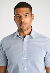 Camicetta a maniche corte con bottoni di colore azzurro chiaro e con un piccolo motivo geometrico. Presenta un colletto a punta e bottoni bianchi a contrasto.