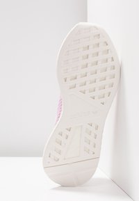 Podešev bílé tenisky Adidas s geometrickým vzorem se opírá o bílou zeď na bílé ploše.