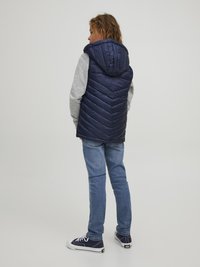 Námornícka puffer vesta s kapucňou, s šípovým prešívaním, ľahký materiál, v kombinácii so šedým dlhým rukávom a modrými džínmi, so športovými teniskami.
