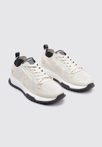 Zapatillas de deporte de color beige claro con parte superior de malla y sintético, media suela blanca, suela negra y sutiles acentos en contraste. Presenta un diseño de cordones.