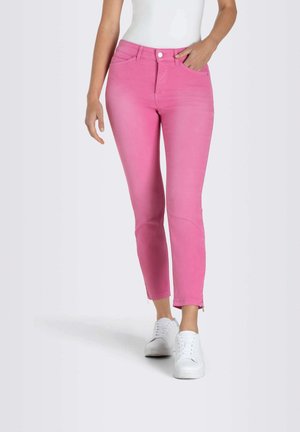 Mujer que lleva puestos pantalones ajustados de color rosa brillante, zapatillas blancas y una camiseta blanca, de pie con una mano en el bolsillo contra un fondo liso.