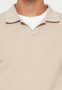 Beige geribbelde polo met een platte kraag en een v-hals, gedragen over een wit t-shirt, met subtiele textuurdetails.