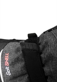 Schwarzes Softshell-Material mit strukturiertem grauen Stoff, ausgestattet mit einem verstellbaren Tragegurt und einem kleinen schwarzen Kunststoffverschluss. Rotes "Soft SHELL"-Logo sichtbar.