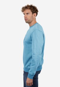 Sudadera azul claro con cuello redondo, puños y borde acanalados. Presenta un diseño sutil en la parte frontal. La tela tiene una apariencia suave y texturizada.