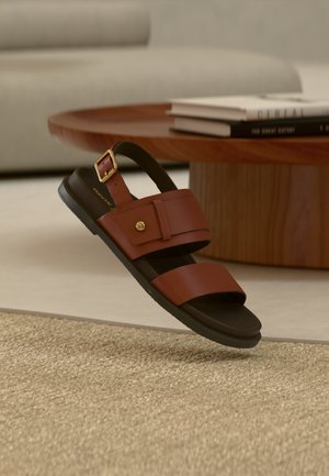 Sandale en cuir marron avec une sangle à boucle flottant au-dessus d'un tapis beige, avec une table en bois et des livres empilés en arrière-plan.