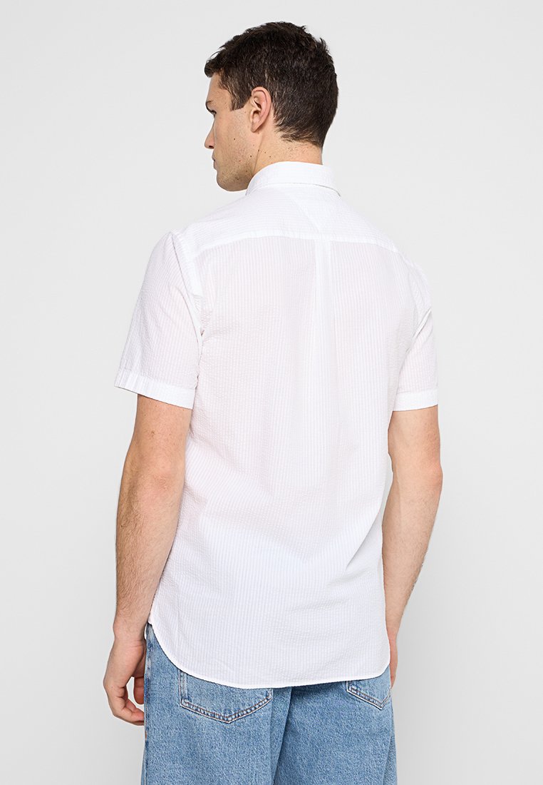 Jeune homme de dos portant une chemise blanche à manches courtes avec des rayures verticales subtiles et un jean en denim bleu clair.