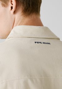 Beige corduroy shirt met een brede kraag en ribbelstructuur, met daarop "PEPE JEANS" geborduurd in marineblauw op de achterkant.