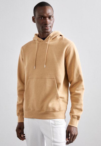 Model masculin îmbrăcat cu un hanorac bej cu glugă și buzunar frontal, purtând pantaloni albi cu logo embosat pe coapsă, stând pe un fundal simplu.