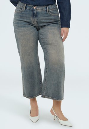 Jeans a gamba larga corti di colore blu sbiadito abbinati a décolleté bianche con cinturino posteriore e camicia di denim scuro a maniche lunghe.