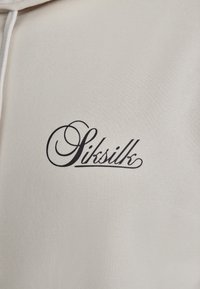 Krém színű kapucnis pulóver, fekete hímzett "Siksilk" logóval, stilizált betűtípussal a mellkas területén. Puha, texturált anyag.