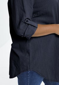 Chemise texturée bleu foncé avec des manches retroussées et un ourlet arrondi ; présentée sur le côté d'un mannequin, associée à un jean bleu.