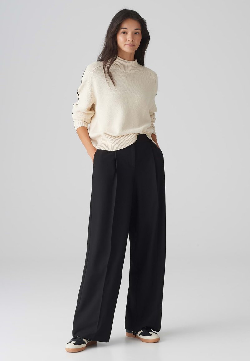 Cremfarbener Strickpullover mit hohem Kragen, kombiniert mit schwarzen weiten Hosen. Schwarze und weiße Sneakers runden das Outfit ab.