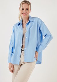 Light blue button-up shirt made of cotton, featuring a classic collar, long sleeves, and a relaxed fit, paired with beige pants.