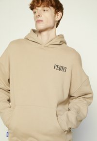 Beige hoodie ar kengura kabatu, ribotiem aproces un logotipu ar uzrakstu "PEQUS" uz krūšu daļas. Izgatavots no mīksta, strukturēta auduma.