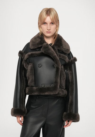 Donna bionda che indossa una giacca in pelle nera con bordo in pelliccia scura e pantaloni in pelle nera, in piedi davanti a uno sfondo bianco uniforme.