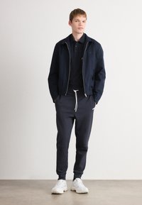 Ung mand stående med hænderne i lommerne på mørke joggingbukser, iført en marineblå zipjakke, sort polo-shirt og hvide sneakers foran en ensfarvet baggrund.