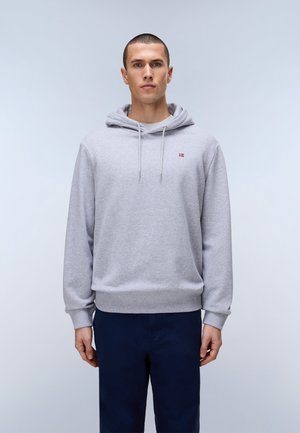 Mann trägt hellgrauen Hoodie mit kleinem Flaggenemblem auf der Brust und dunkelblaue Hose, steht vor einfarbig hellem Hintergrund.