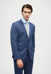 Νεαρός άνδρας φορώντας μπλε καρό κοστούμι, ανοιχτό μπλε πουκάμισο και μπεζ γραβάτα, standing against a plain white background.