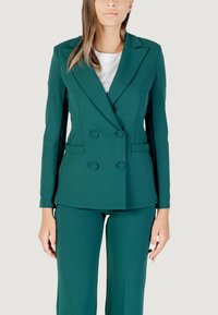 Modelo vistiendo una chaqueta de traje verde ajustada sobre una blusa blanca, combinada con pantalones a juego, sonriendo frente a un fondo neutro.