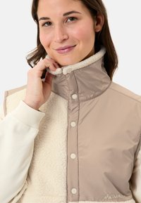 Gilet rembourré beige et crème avec un col montant, doté d'une façade en polaire texturée, de boutons-pression et de poignets en tricot côtelé.