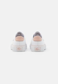 White sneakers with pink heel accents, textured rubber outsole, and a logo detail on the heel. Smooth material with stitched accents.