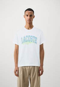 Balta kokvilnas T-kreklā ir zils un zaļš uzraksts "LACOSTE" ar krokodila grafiku, apaļa kakla izgriezums un īsas piedurknes.