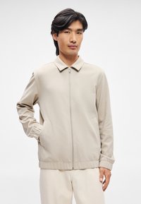 Jasno beżowa kurtka zapinana na zamek z kołnierzem, długimi rękawami, bocznymi kieszeniami i elastycznymi mankietami. Gładki materiał o czystym, minimalistycznym wzornictwie.