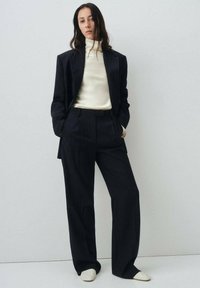Blazer noir à rayures fines sur mesure et pantalon évasé associés à un haut crème à col montant et des chaussures de couleur claire sur un fond neutre.