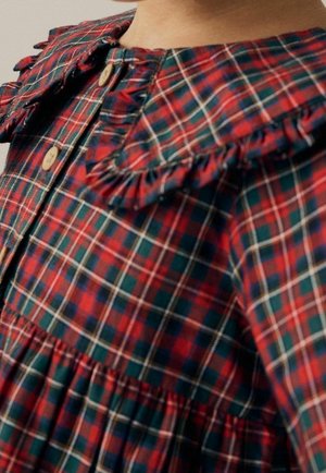 Enfant portant une robe à carreaux rouge et vert avec des boutons en bois et des volants près du col et de la taille.