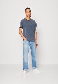 Una camiseta de manga corta azul con un aspecto desgastado, combinada con jeans de lavado claro y zapatillas blancas. La camiseta presenta un pequeño logotipo.