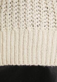 Chunky gestrickter cremefarbener Pullover mit ribbeliger Textur am Saum, die sich von einem glatten schwarzen Stoff abhebt. Verfügt über offene Stiche.