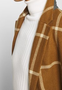 Cappotto a quadri marrone con accenti beige, indossato sopra un dolcevita bianco a coste. Il cappotto presenta linee sulle spalle ben definite e un colletto classico.