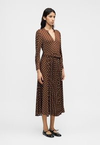 Καφέ πουά φόρεμα με μακριά μανίκια, V-λαιμόκοψη και ζώνη στη μέση. Ροή φούστα με πτυχώσεις συνδυασμένη με μαύρα επίπεδα παπούτσια.