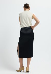 Frau mit einem Dutt trägt ein ärmelloses, elfenbeinfarbenes TextTop, einen schwarzen Midikleid Stiftrock mit seitlichem Schlitz und beigen geflochtenen High Heels und steht mit einer Hand auf der Hüfte.