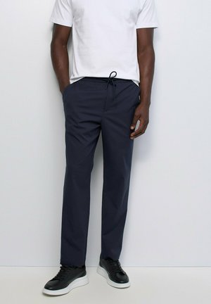 Hombre que lleva pantalones azul marino con cordón, camisa blanca de manga corta y zapatillas negras con suelas blancas, de pie contra un fondo blanco liso.