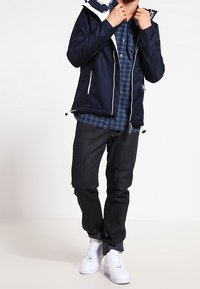 Giacca impermeabile blu navy con accenti bianchi, sopra una camicia a quadri blu. Pantaloni scuri e sneakers bianche completano l'outfit.