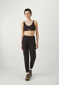 Črni športni modrček z izrezi, v kombinaciji s črnimi športnimi hlačami. Model nosi rjave pohodniške čevlje in bele nogavice. Nevtralna podlaga.