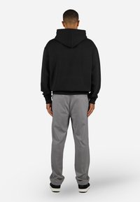 Mand står med ryggen til iført en sort hættebluse, grå bukser og sort-hvide sneakers på en hvid baggrund.