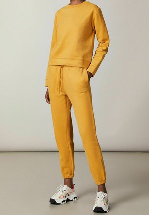 Gele sweatshirt met bijpassende joggingbroek van zachte stof, voorzien van een ronde hals, geribbelde boorden en een elastische tailleband. Geaccentueerd met minimale branding.