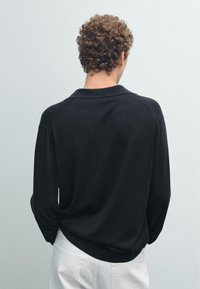 Černý pletený svetr s uvolněným střihem, dlouhými rukávy a kulatým límcem, kombinovaný se světlými kalhotami. Viditelný strukturovaný detail látky.