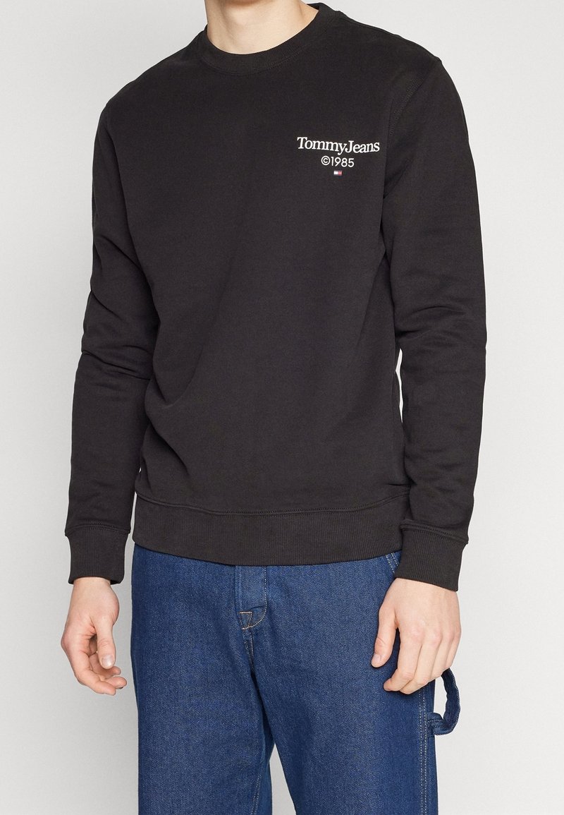 Personne portant un sweat-shirt noir Tommy Jeans avec le texte 1985 et un jean bleu, debout devant un fond clair uni.