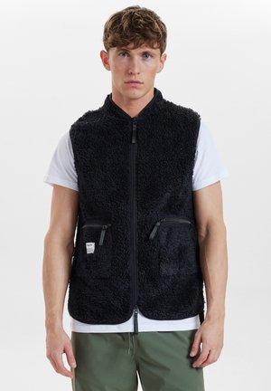 Chaleco negro forrado de felpa con cierre de cremallera y dos bolsillos frontales con cremallera. Usado sobre una camiseta blanca, con una tela texturizada y despeluchada.