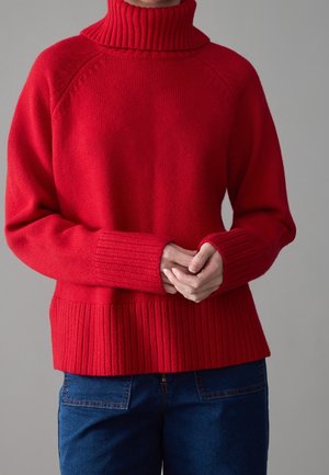 Personne portant un pull col roulé rouge vif avec poignets et ourlet côtelés, associé à un jean bleu foncé, mains jointes devant.